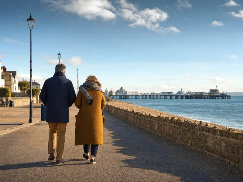 Two people in warm coats walking side by side along the Southsea seafront promenade on a bright morning, with the Solent and a pier stretching out in the distance.